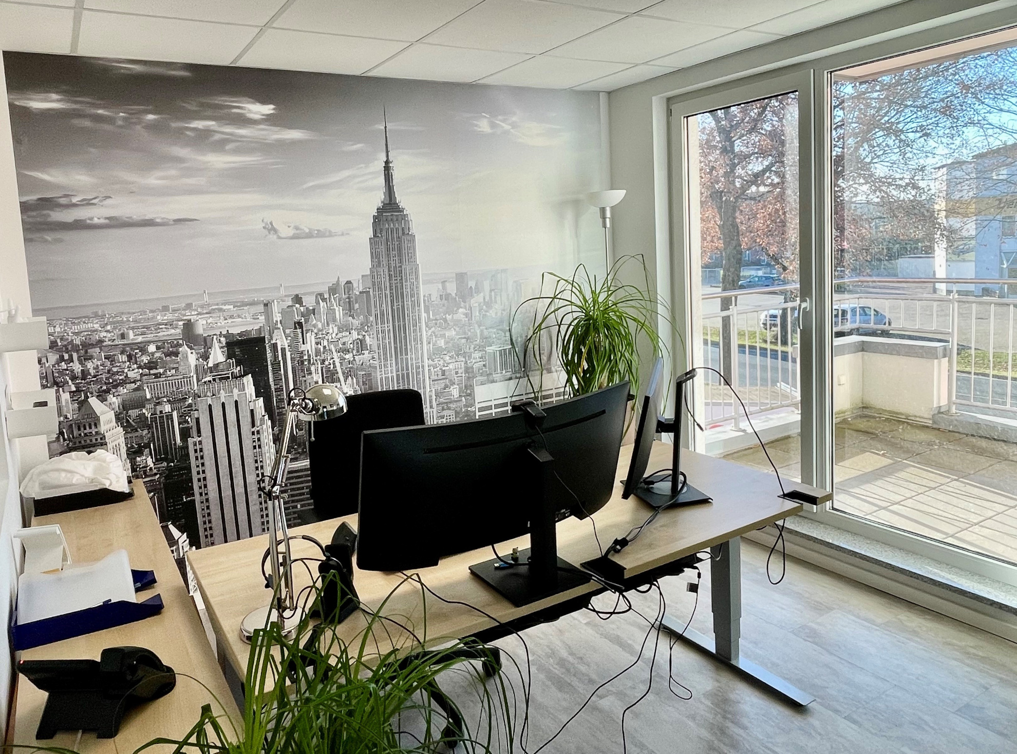 Ein lichdurchflutetes Einzelbüro mit Grünpflanzen und Balkonzugang. Im Zentrum ein großer Eckschreibtisch, ausgestattet mit 2 Monitoren, vor der Skyline von New York.