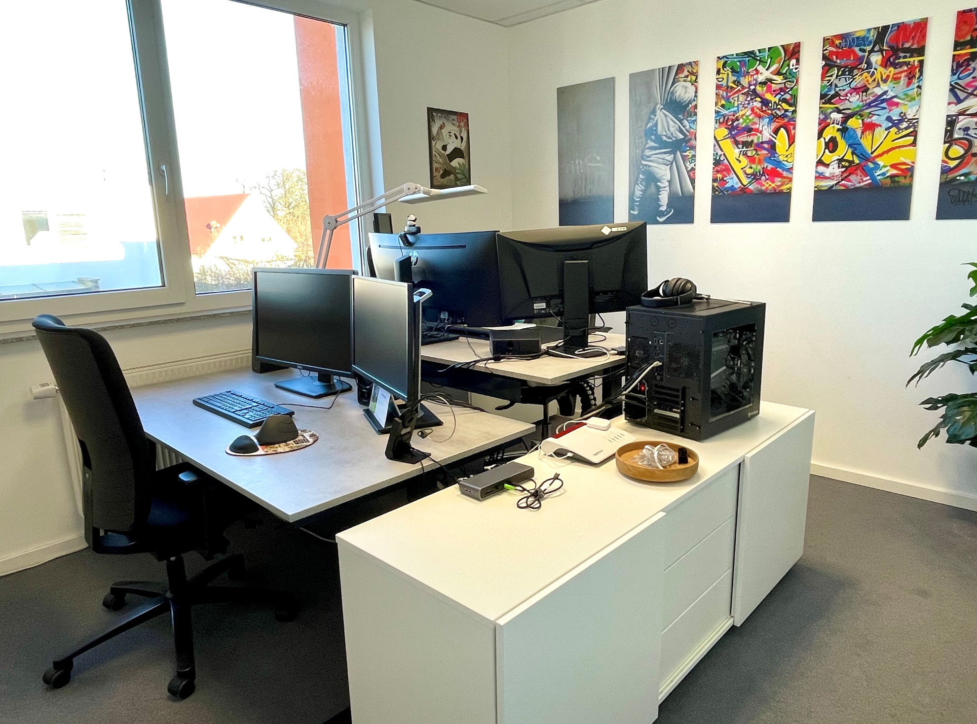 Ein helles 2-Personen-Büro mit großem Fenster, ausgestattet mit 2 Schreitischen, mit je 2 Monitoren, und einem schreibtischhohen, langen Material-Schrank. Im Hintergrund sind 5 farbenfrohe, längliche Bilder an der langen Wand geradezu, angebracht und rech